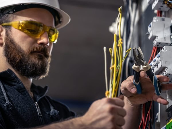 A male electrician works in a switchboard with an electrical connecting cable, connects the equipment with tools.